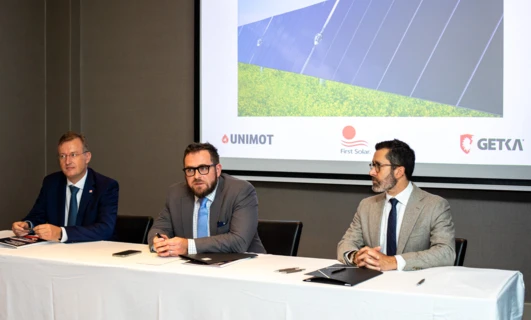 Pracownik biurowy w eleganckim ubraniu trzymający mikrofon na wydarzeniu z ubraniami, odzieżą zewnętrzną i urządzeniami elektronicznymi. Logo UNIMOT, GETKA i First Solar widoczne w widocznym miejscu.