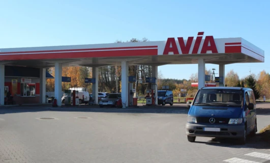 Samochód rodzinny na stacji benzynowej tankujący benzynę. Widoczne są zderzak, lusterka i tablica rejestracyjna pojazdu. Tablice informacyjne wskazują na lokalizację jako KIOPIST Sklep ON AVIA 94.