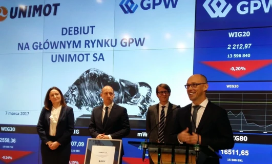 Tekst alternatywny: Profesjonalista w formalnym stroju białego kołnierza z płaszczem, marynarką i logo UNIMOT. Dane giełdowe w języku polskim na Giełdzie Papierów Wartościowych w Warszawie.