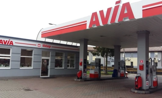Zbliżenie dystrybutora paliwa na stacji benzynowej AVIA z lustrem samochodowym, zderzakiem i tablicą rejestracyjną w tle. - Stacja benzynowa AVIA.